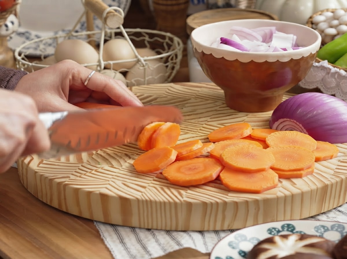 나무 도마 위에서 껍질 벗긴 당근을 둥근 동전 모양으로 썰고 있는 손.