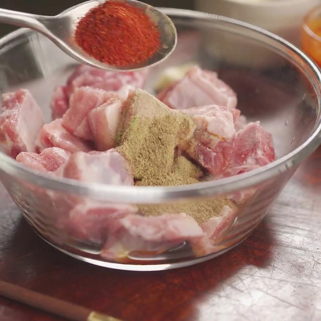 혼합 향신료와 허브로 덮인 생돼지 갈비 볼 위로 숟가락에 담긴 붉은색 칠리 가루가 보이는 모습.