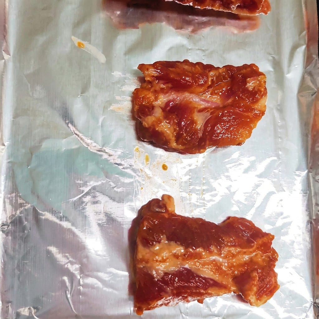 알루미늄 호일이 깔린 베이킹 시트에 한 줄로 나란히 정렬된 양념된 돼지고기 갈비 조각들.