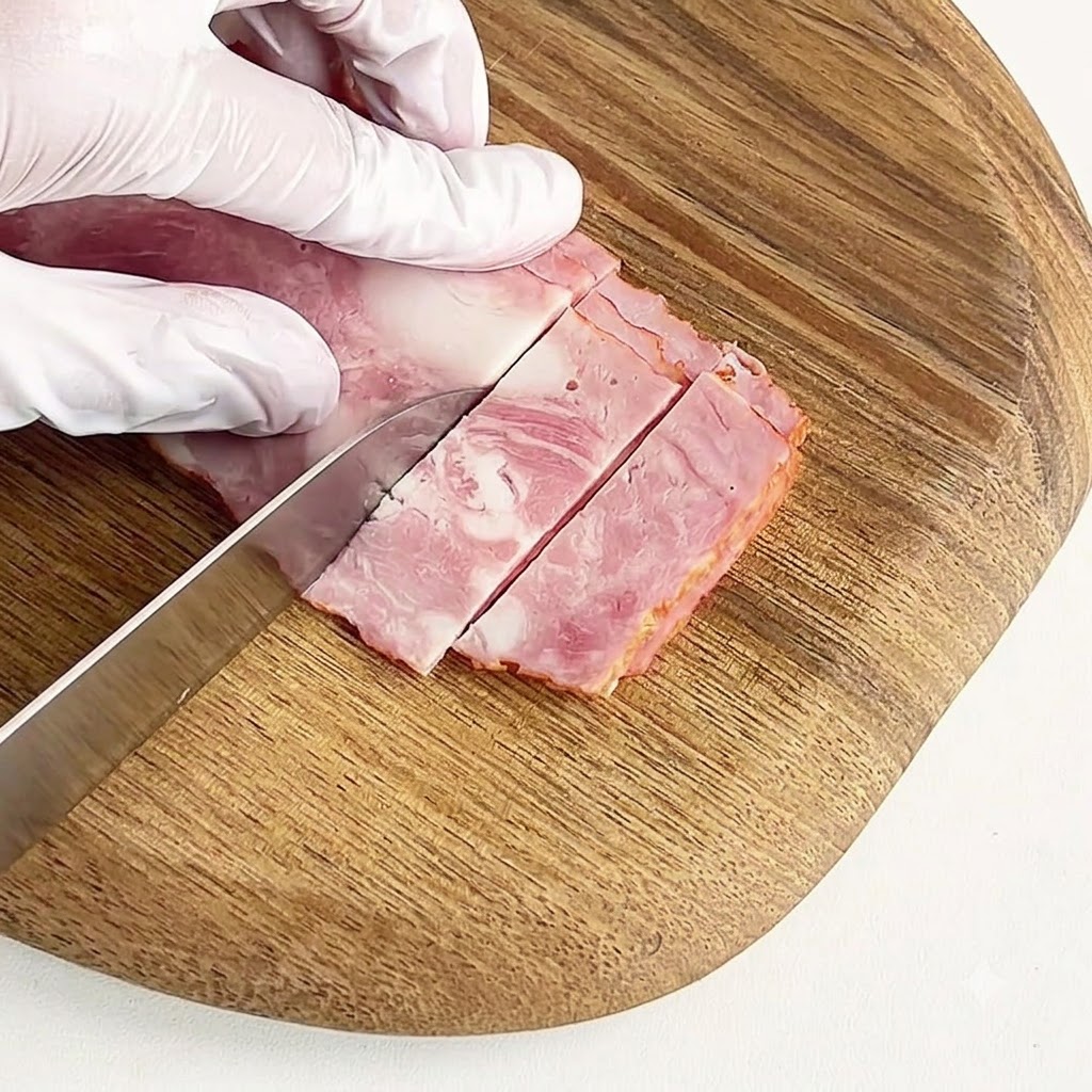 장갑을 낀 손이 둥근 나무 도마 위에서 숙성된 베이컨을 작은 조각으로 썰고 있는 모습.