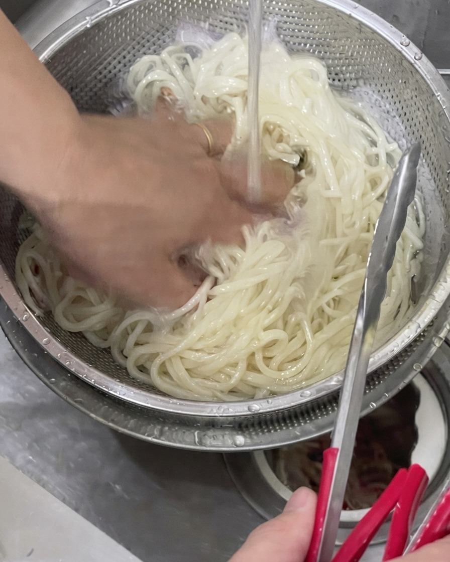 차가운 수돗물이 흐르는 금속 체에 걸러진 삶은 우동면을 손으로 헹구는 모습
