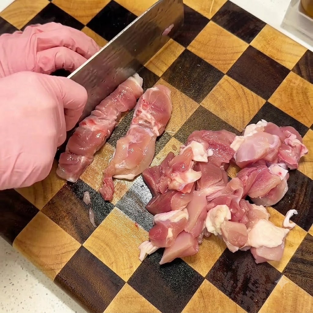 핑크색 장갑을 낀 손으로 도마 위에서 생닭다리살을 깍둑썰기하는 모습.