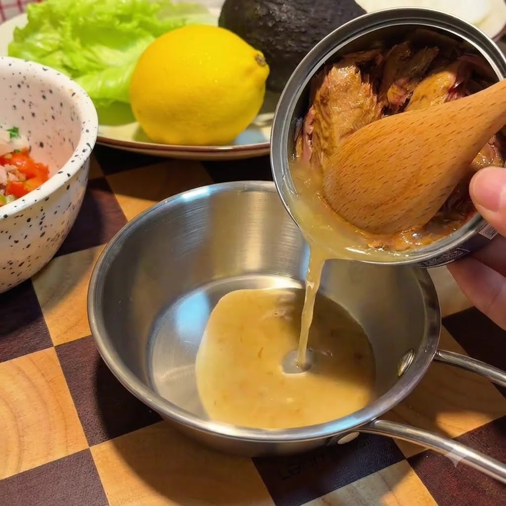 나무 숟가락으로 캔 안의 참치 살을 누르며 황금빛 오일을 작은 스테인리스 그릇에 따르는 모습.