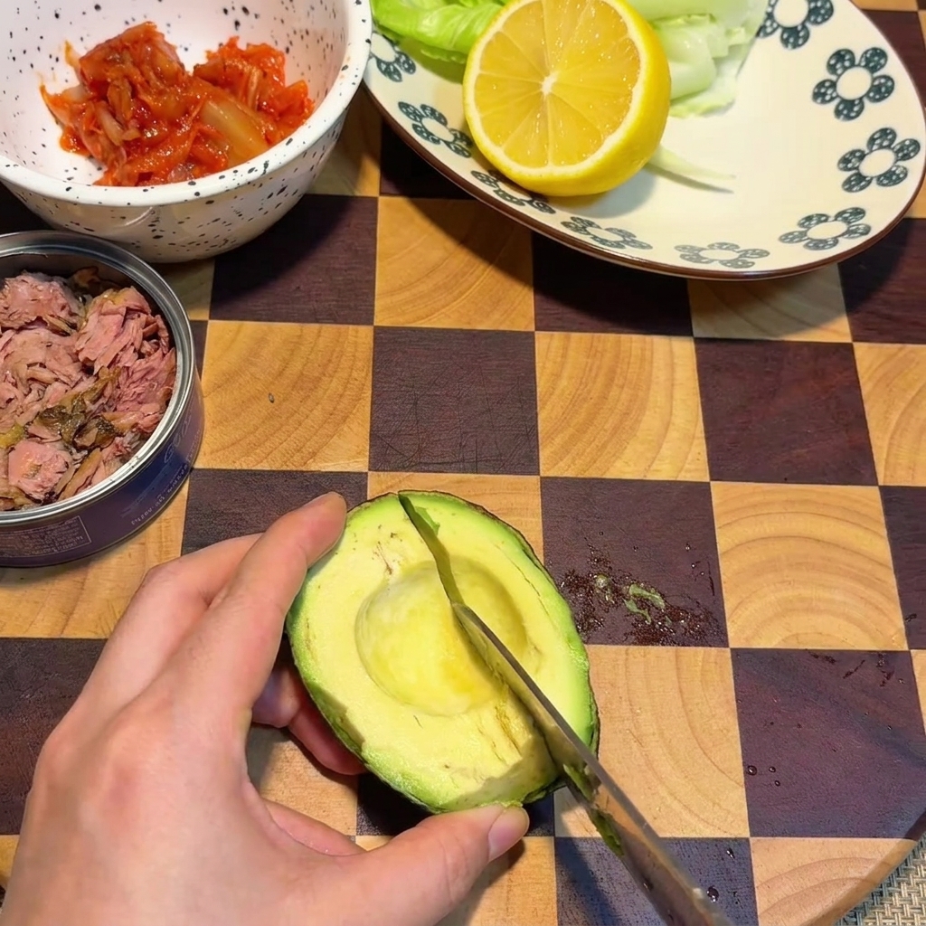 격자무늬 나무 도마 위에 놓인 아보카도 반쪽의 과육에 칼로 격자 모양의 칼집을 내는 손.