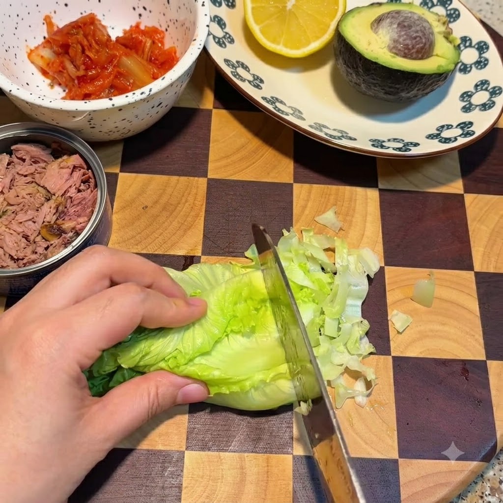 김치 그릇과 참치캔 옆 격자무늬 도마 위에서 신선한 초록색 양상추 잎을 칼로 써는 모습.