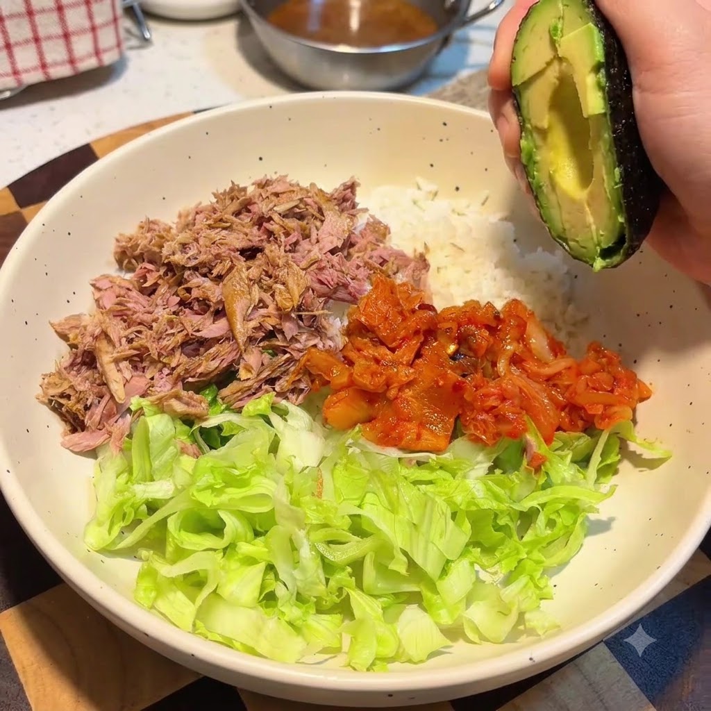 밥, 양상추, 참치, 김치가 담긴 큰 그릇 위에 아보카도 반쪽을 들고 있는 손.