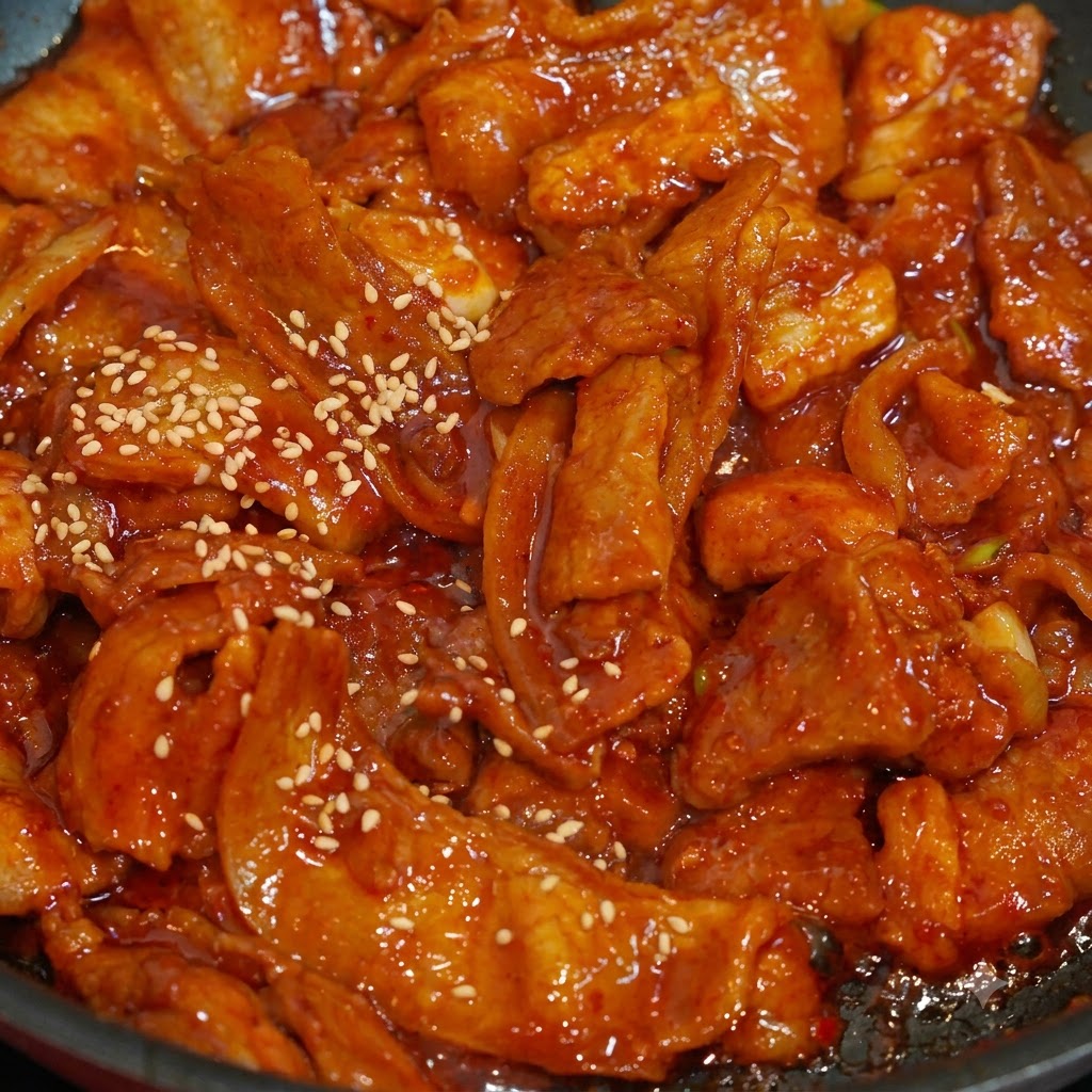 팬 안의 빨갛고 먹음직스러운 매운 제육 위에 흰 참깨가 뿌려지는 클로즈업 샷.