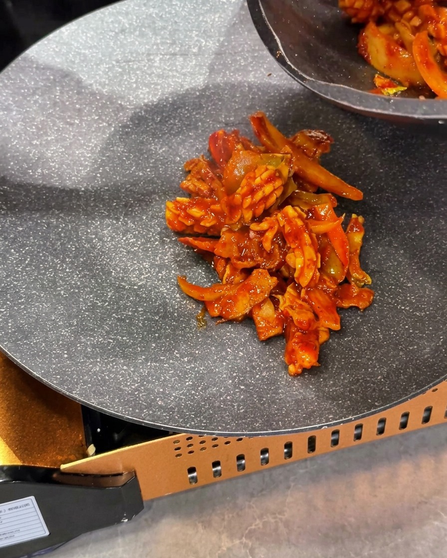 조리된 붉은색 오징어 및 삼겹살 볶음 요리를 휴대용 가스 버너 위의 팬 중앙에 담고 있는 모습