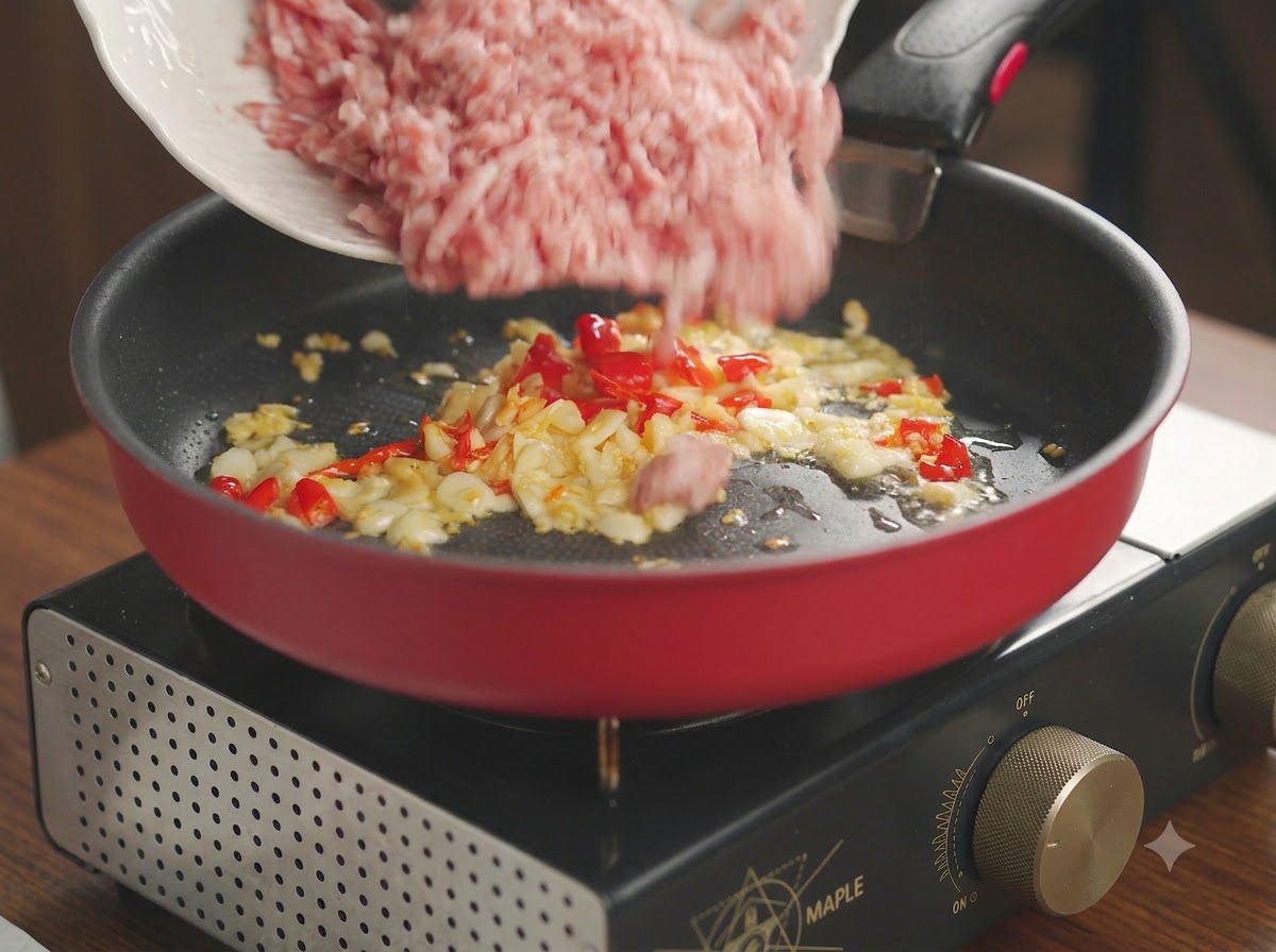 Raw minced pork being poured from a white plate into a red frying pan with fried garlic and chili.