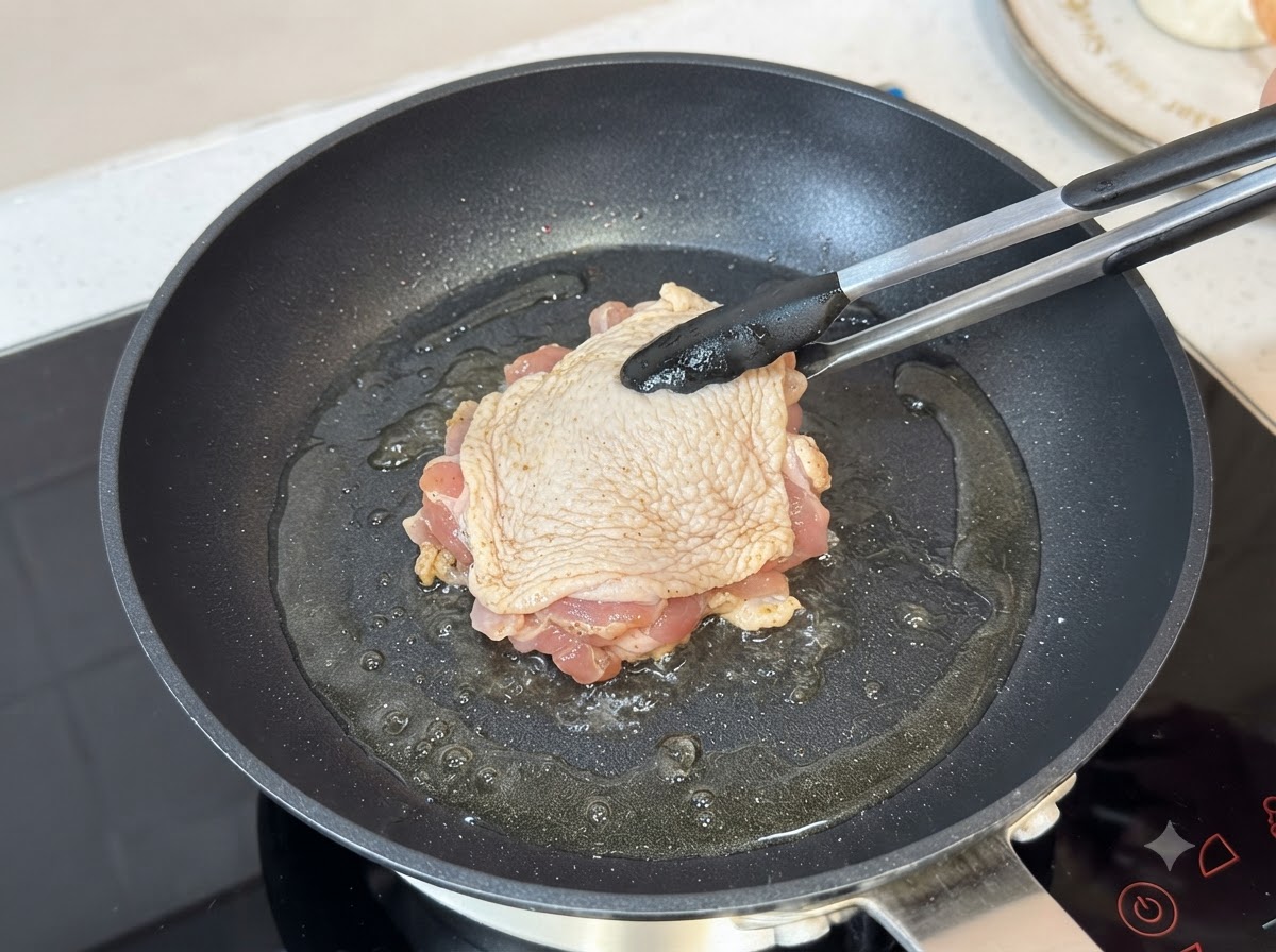 기름을 두른 뜨거운 팬에 닭다리살 껍질이 아래로 향하게 들어가 집게로 집혀 있는 모습.
