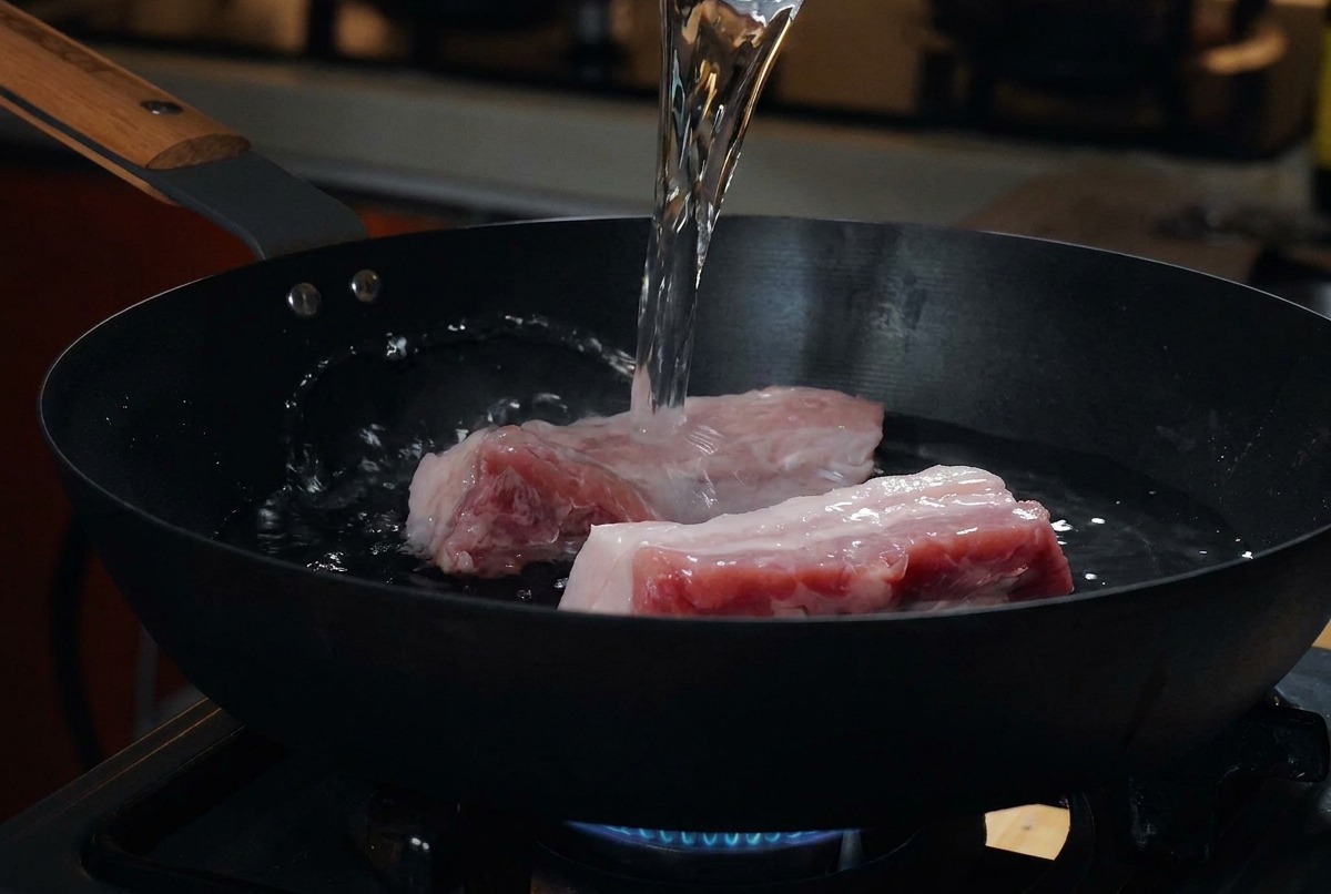 차가운 검은색 팬에 생삼겹살을 올리고 계량컵으로 물을 붓는 모습.