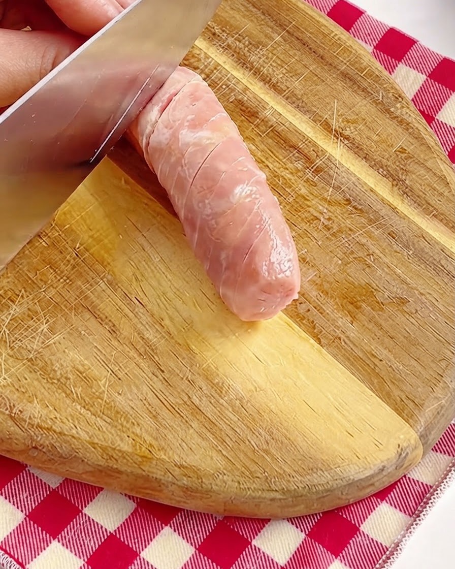 나무 도마 위에서 손으로 칼을 이용해 통통한 분홍색 소시지 표면을 조심스럽게 칼집 내는 모습.