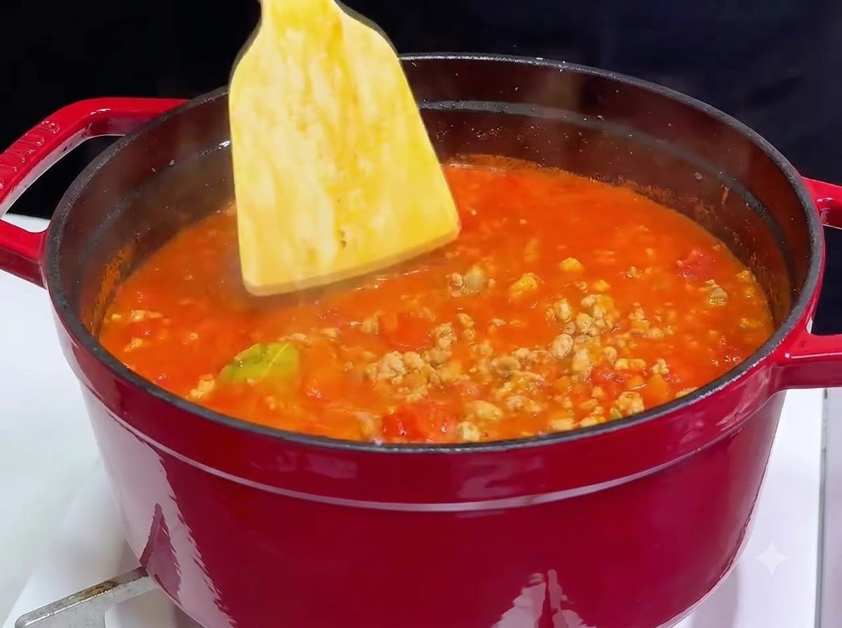 보글보글 끓는 토마토 고기 소스가 가득 찬 빨간색 무쇠 냄비에 나무 주걱으로 저어가며 레드 와인을 섞는 모습.