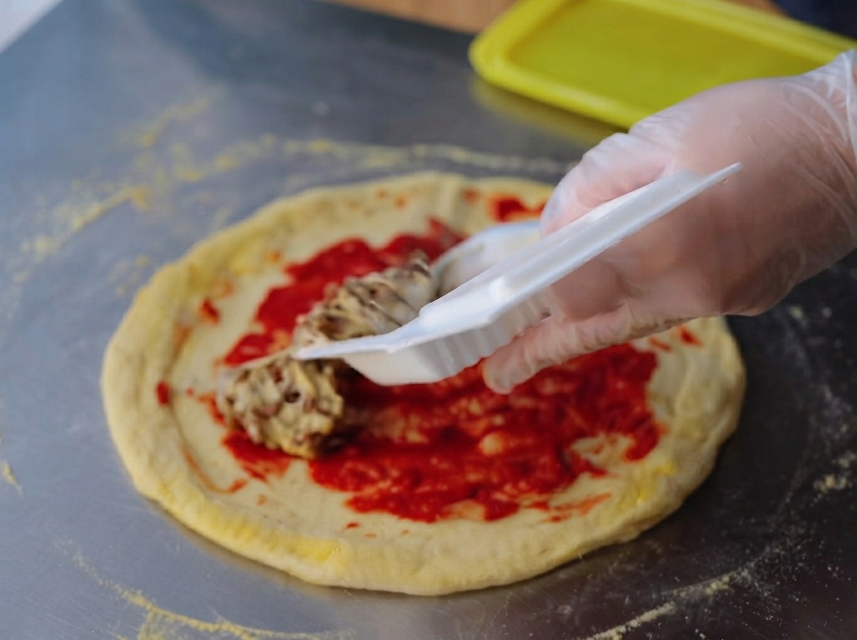 토마토 페이스트를 바른 피자 반죽 위에 끈적이는 낫토를 고르게 펴 올리는 모습.