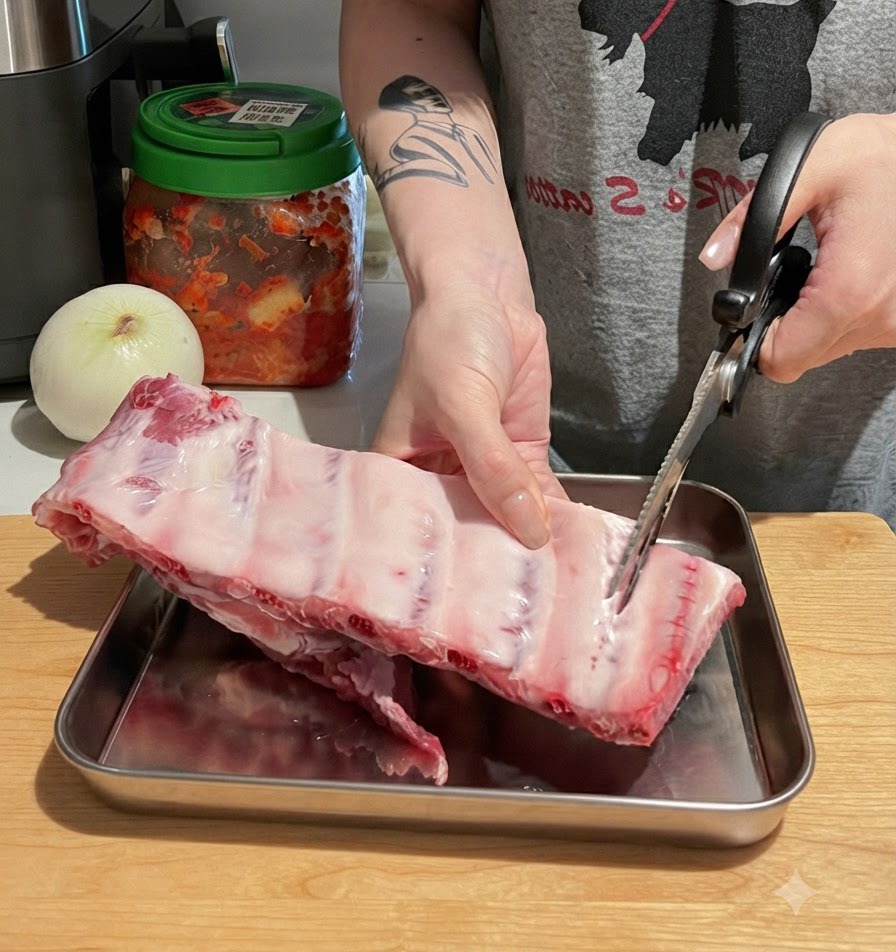 도마 위에서 주방 가사 가위로 커다란 생돼지갈비 뼈대를 한 덩이씩 자르고 있는 손