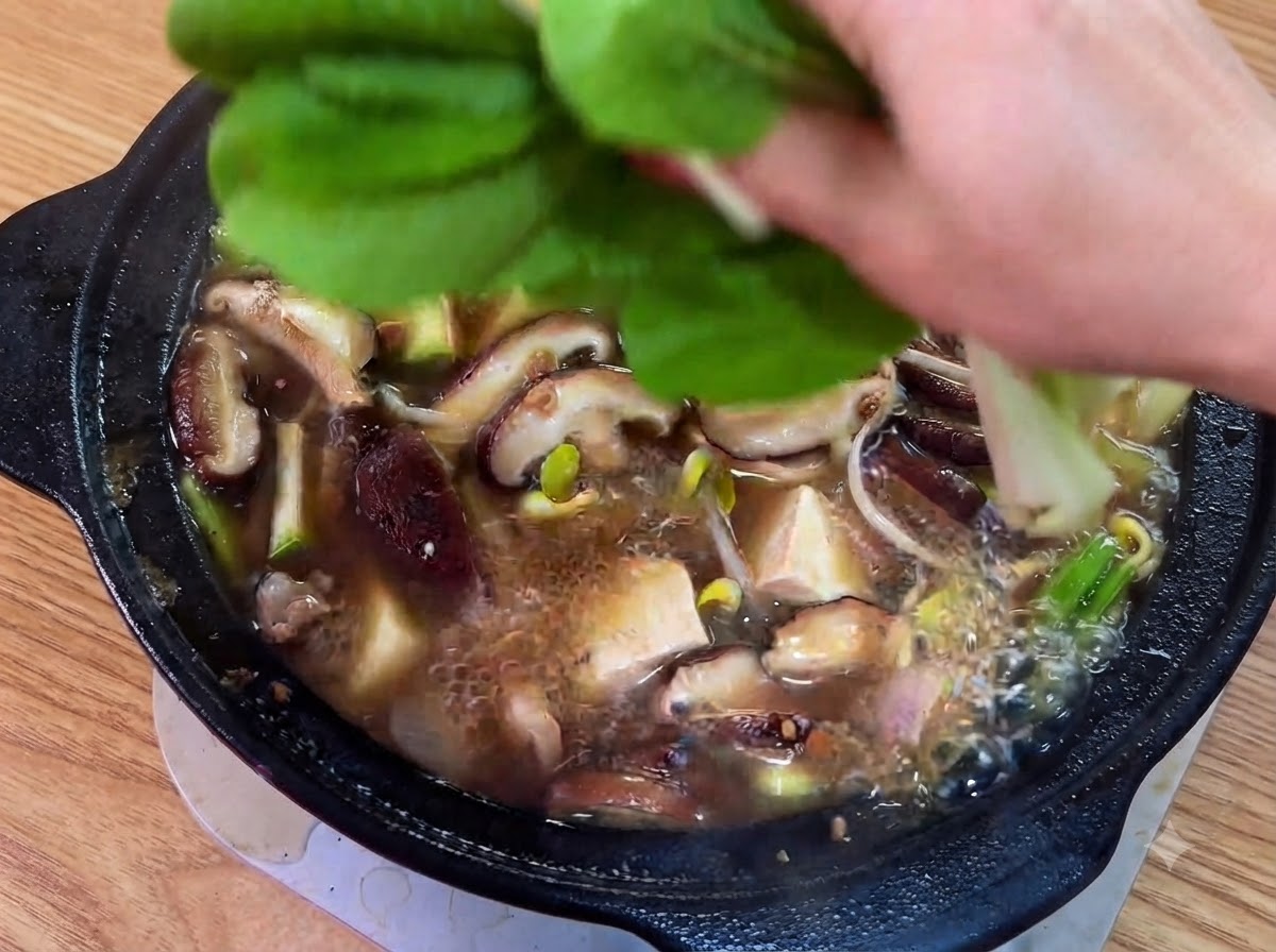 끓고 있는 찌개 위에 신선한 녹색 잎채소와 썬 청고추를 올리는 모습.