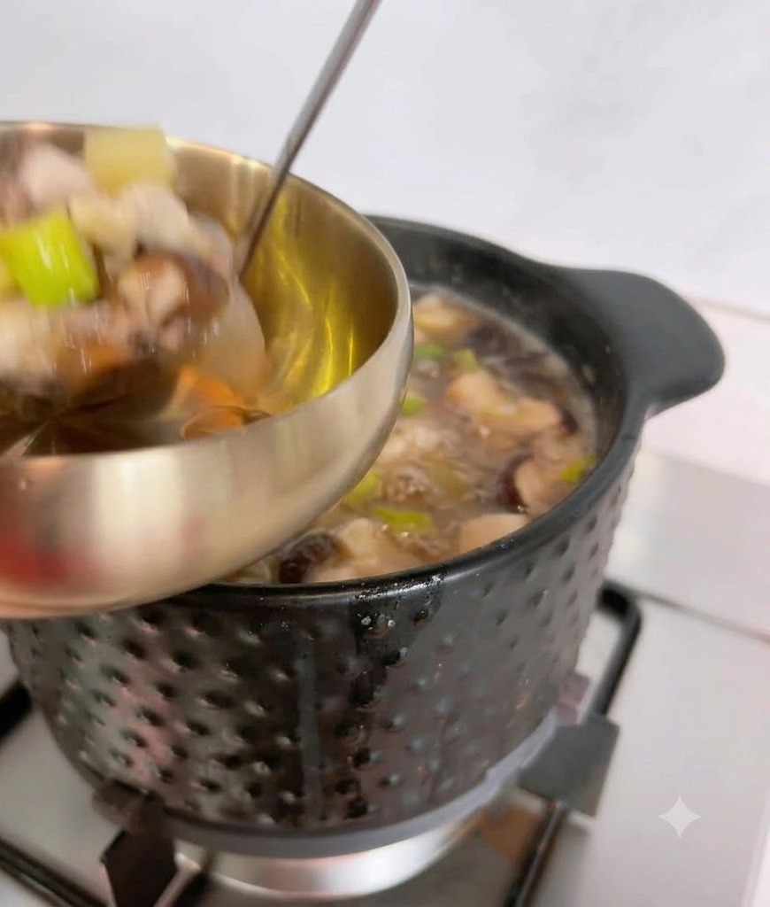 국자로 검은색 냄비에서 뜨거운 된장찌개 국물과 건더기를 퍼서 금속 서빙 그릇에 담는 모습.