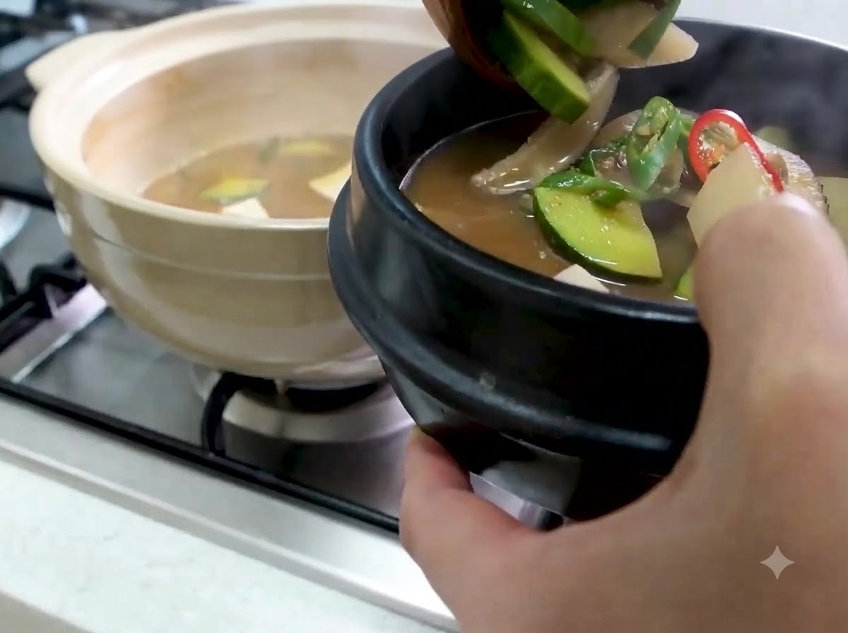 애호박과 채소가 들어간 뜨거운 된장찌개를 국자로 떠서 검은색 돌솥 서빙 그릇에 담는 모습.