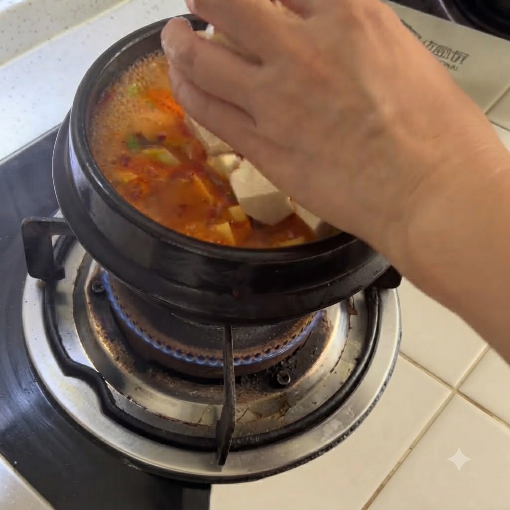 매콤한 국물이 끓는 뚝배기에 손으로 하얀 두부 조각을 넣는 모습.