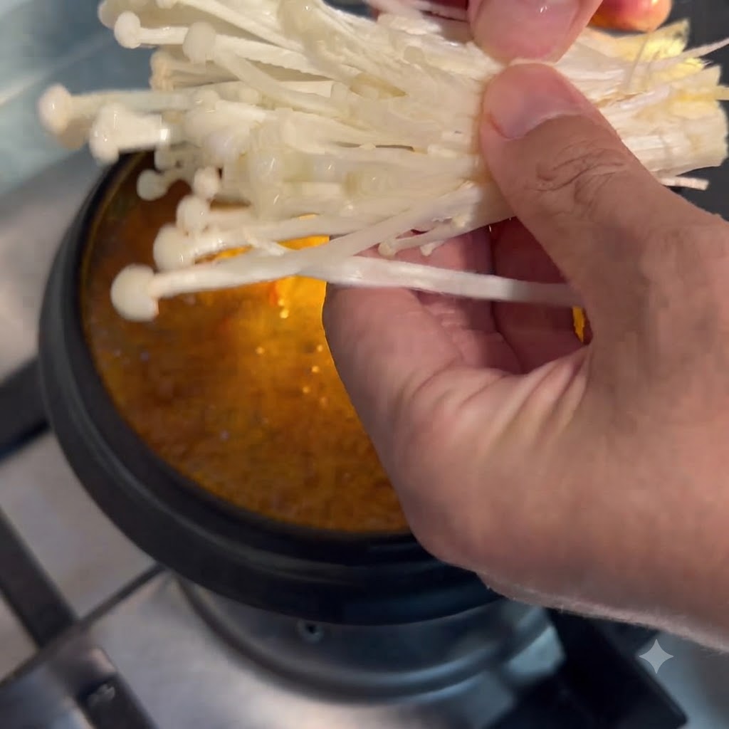 손으로 팽이버섯을 집어 시원하게 끓고 있는 찌개에 넣는 모습.