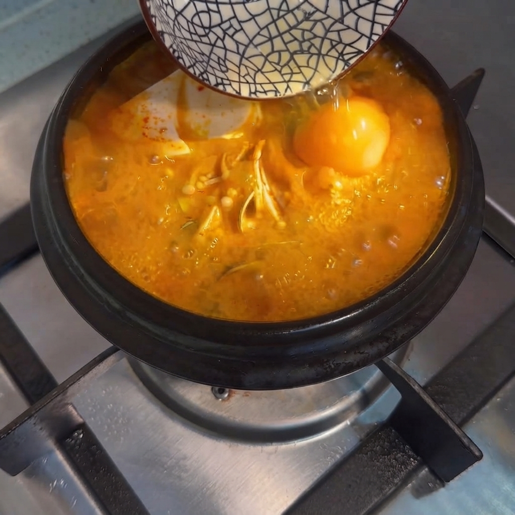 작은 그릇에 담긴 날달걀이 끓는 순두부찌개 중앙으로 떨어지는 모습.