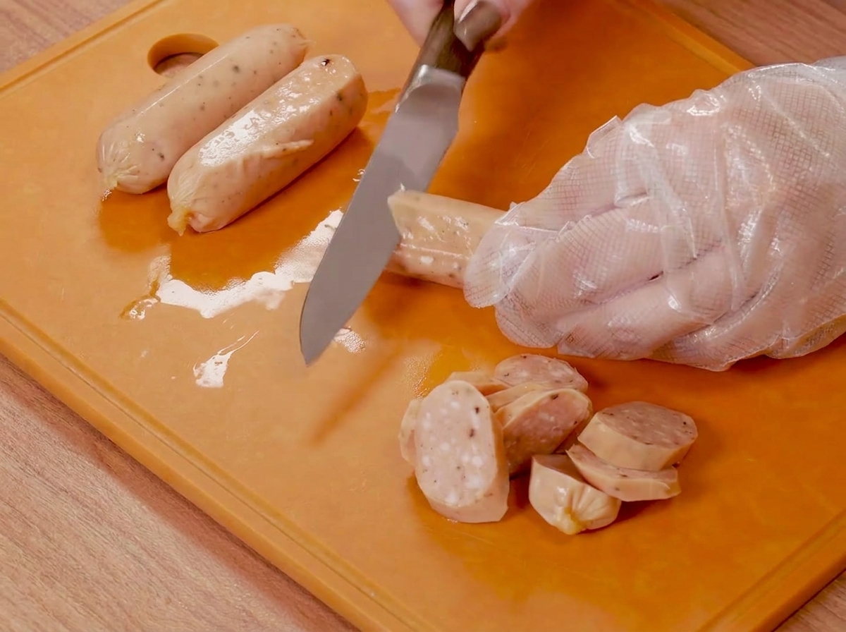 주황색 도마 위에서 장갑을 낀 손이 칼을 이용해 작은 소시지에 칼집을 내고 있는 모습.