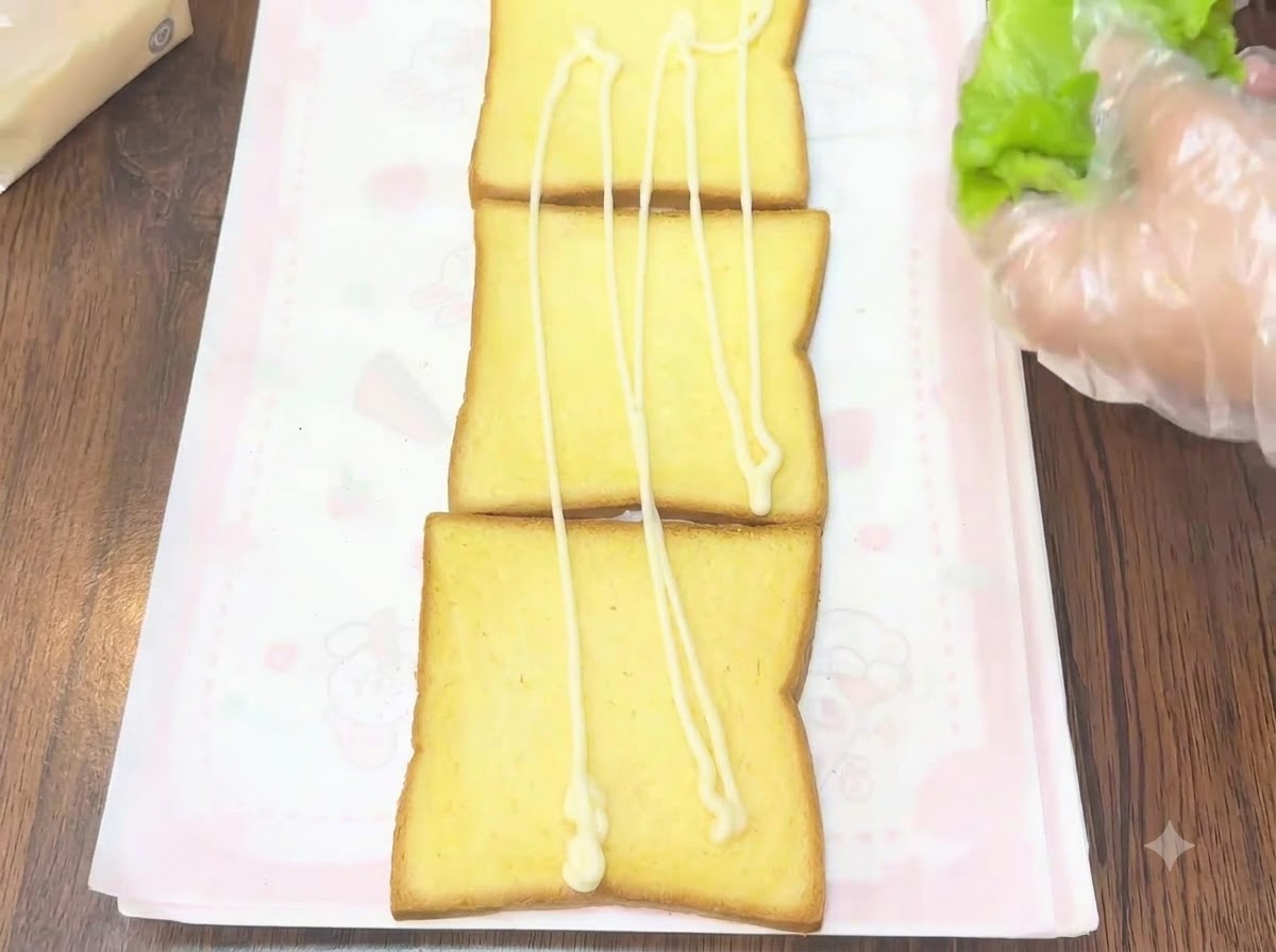 장갑을 낀 손이 드레싱이 뿌려진 식빵 위에 신선한 녹색 상추 잎을 올리는 모습.