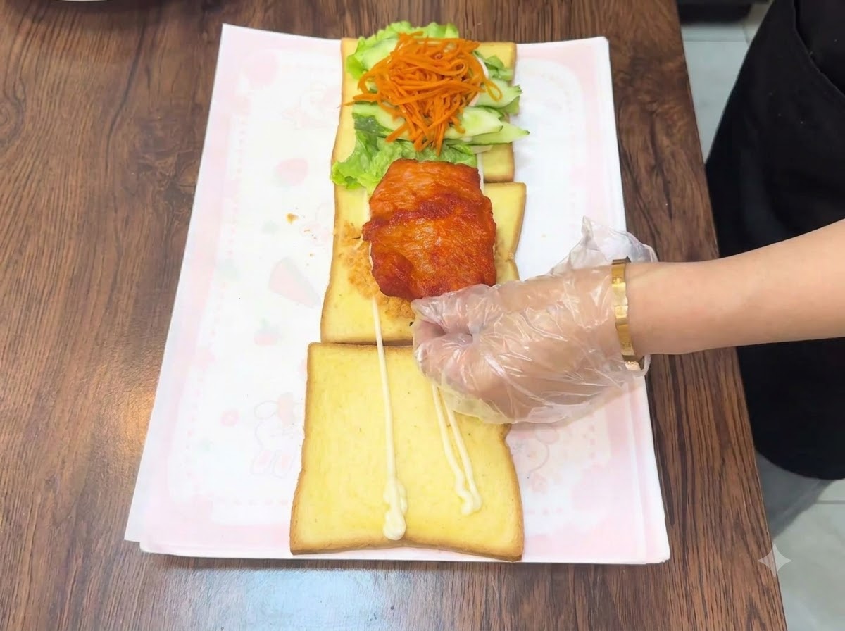 장갑을 낀 손이 층층이 쌓인 토스트 조립품 위에 따뜻한 치킨 커틀릿 튀김을 올리는 모습.