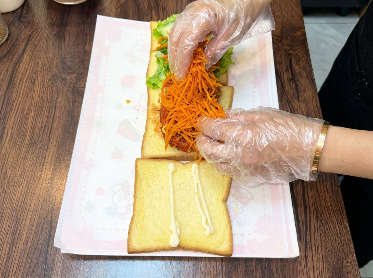 장갑을 낀 양손이 포개어진 치킨 커틀릿 위에 선명한 주황색 볶은 당근채를 듬뿍 쌓아 올리는 모습.