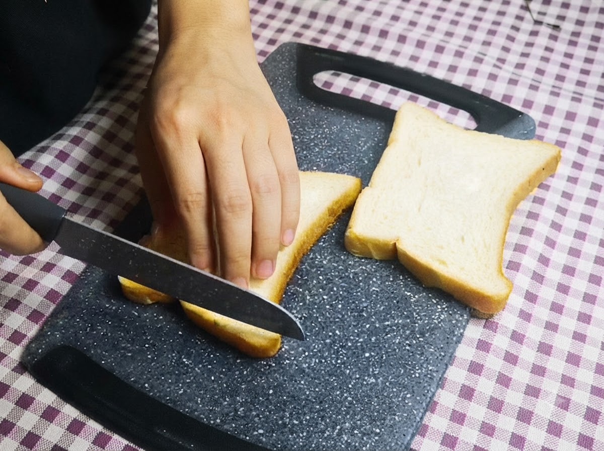 도마 위에서 칼을 사용하여 흰 식빵의 테두리를 자르고 있는 사람의 손.