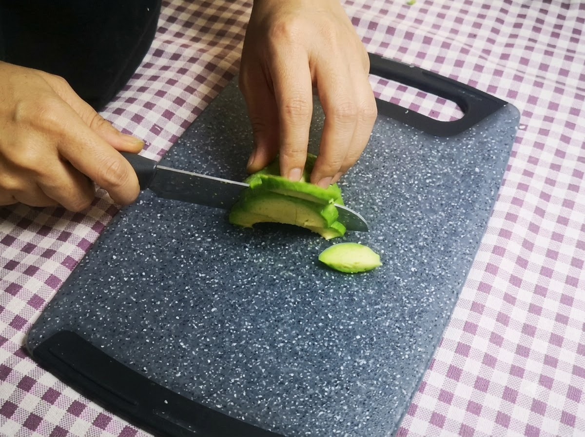 도마 위에서 칼로 껍질을 벗긴 아보카도 반쪽을 얇게 썰고 있는 손.