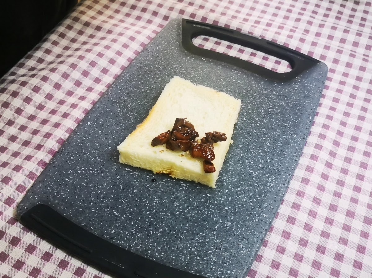 회색 도마 위에 놓인 한 장의 흰 식빵 정중앙에 진한 양념이 벤 닭고기 조각들이 가지런히 놓여 있는 클로즈업 샷.
