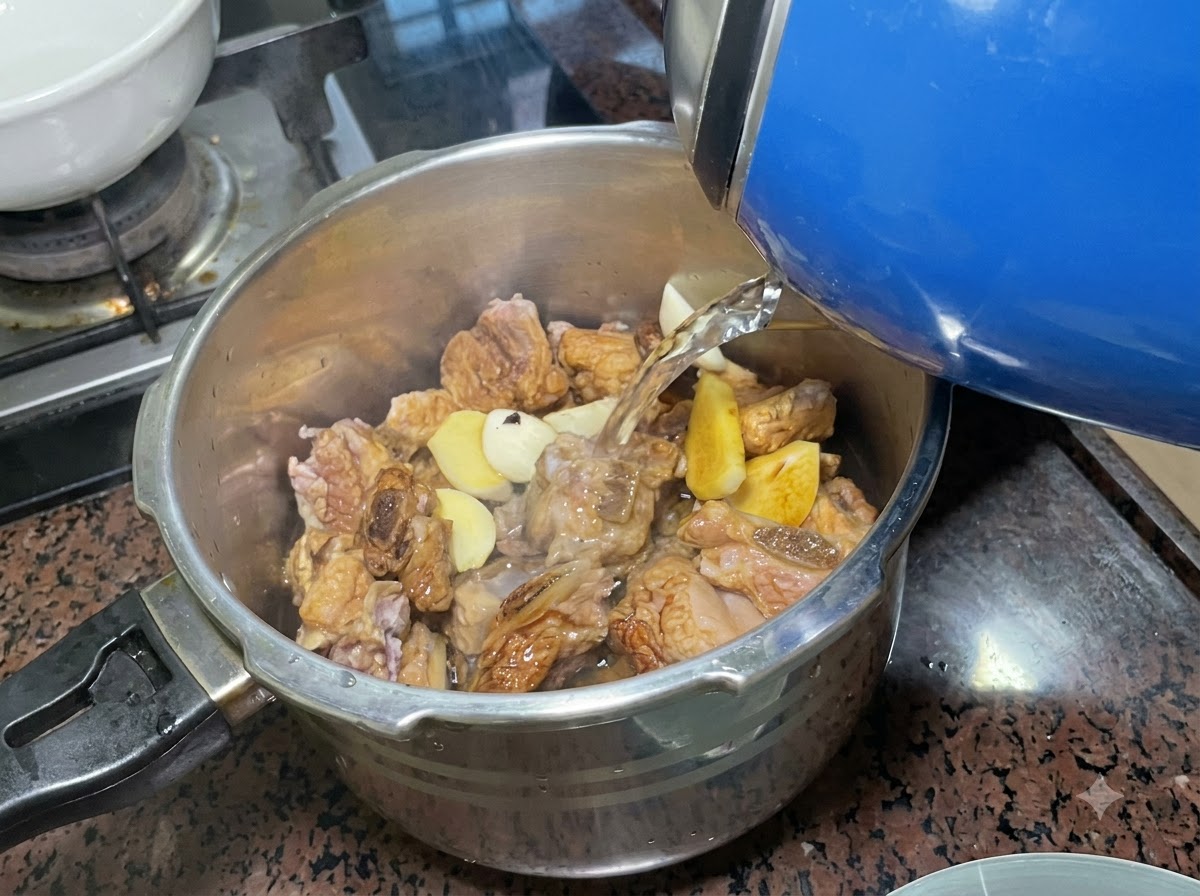 양념된 갈비와 향신채가 채워진 스테인리스 압력솥에 파란색 전기 포트로 물을 붓고 있는 모습.