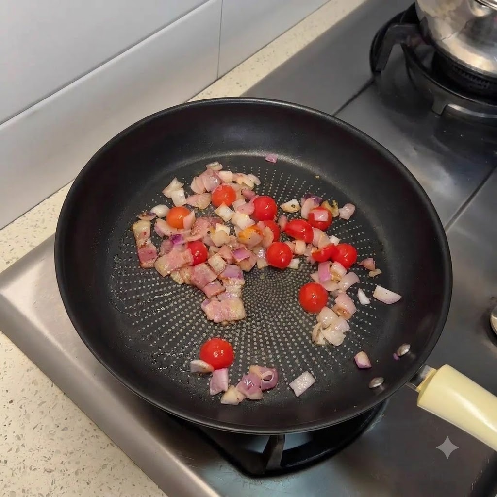 볶은 베이컨, 다진 양파, 반으로 썬 방울토마토가 담겨 있는 프라이팬.