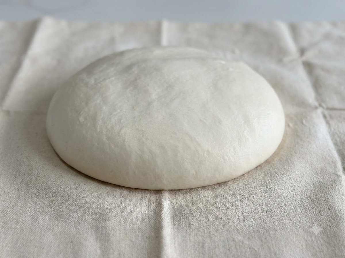 밀가루가 뿌려진 하얀 천 표면 위에 둥글고 매끄러운 흰 반죽 돔이 놓여 있는 모습