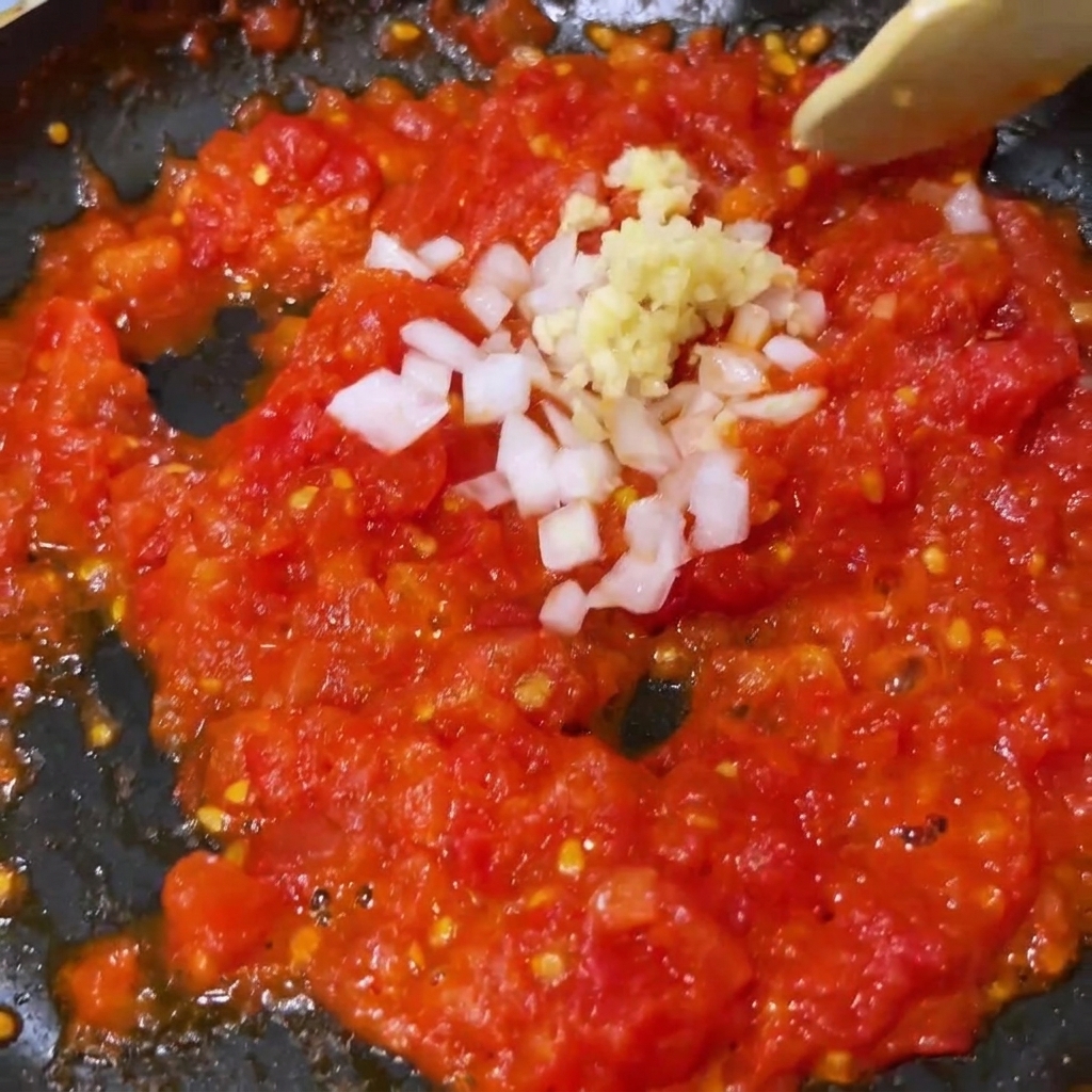 팬에서 부드러워진 으깬 토마토에 다진 마늘과 흰 양파를 섞는 모습.
