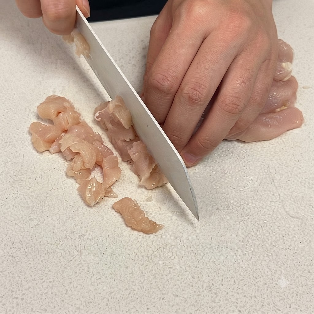 하얀 도마 위에서 날카로운 식칼로 닭가슴살을 작은 큐브 모양으로 썰고 있는 클로즈업 샷.
