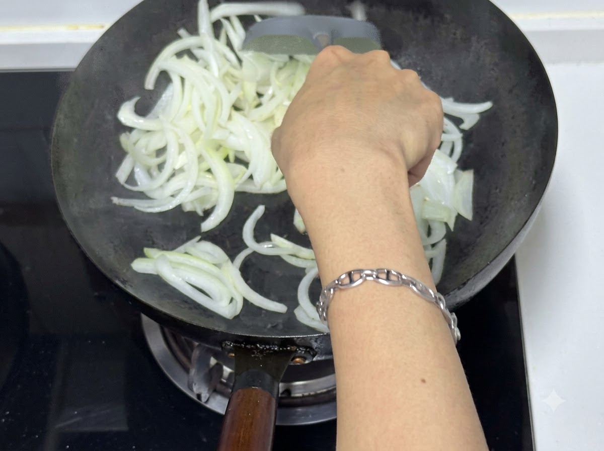 투명해지고 살짝 갈색빛을 띄는 양파를 주걱으로 웍에서 볶고 있는 사람의 모습.