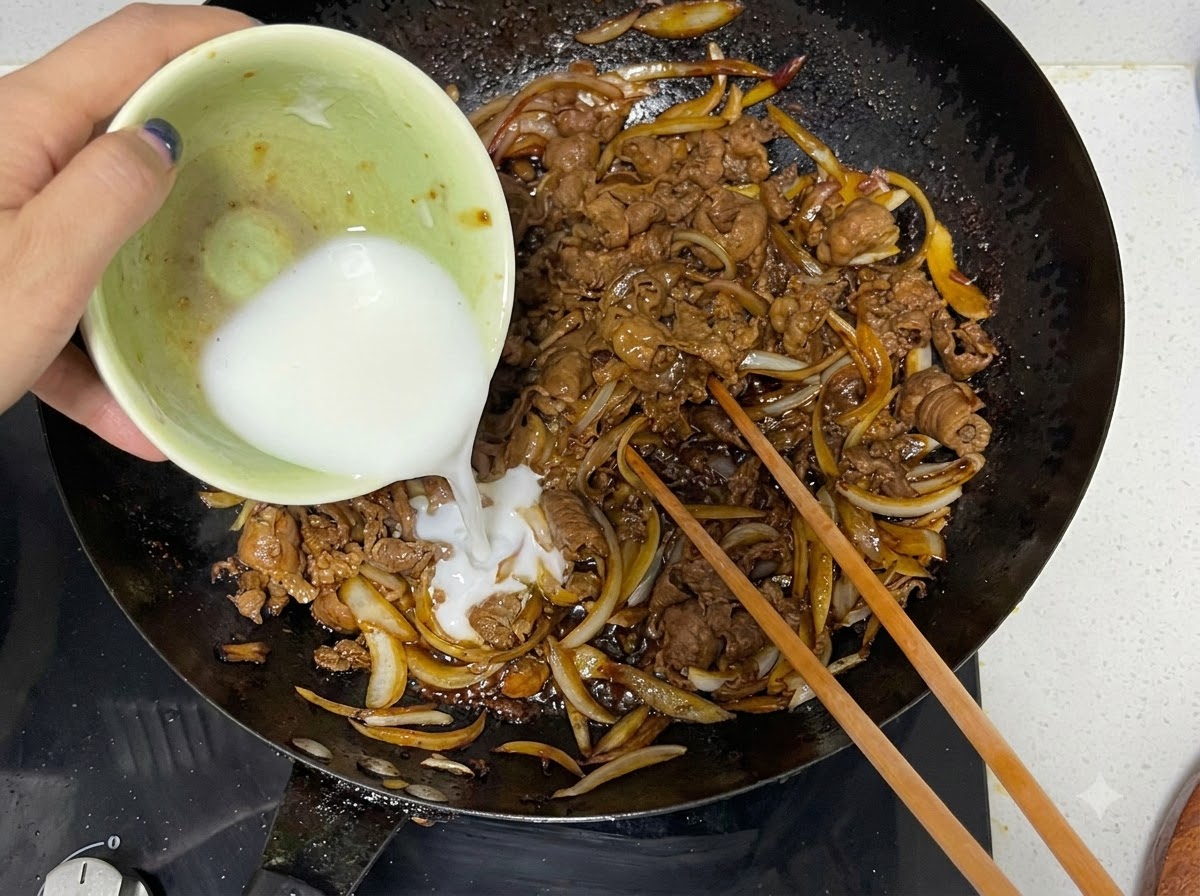 요리용 젓가락과 함께 조리된 소고기와 양파가 담긴 웍에 그릇에서 전분 물이 부어지는 모습.