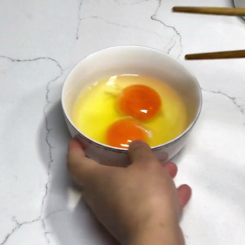 흰 그릇에 담긴 두 개의 계란을 젓가락으로 젓고 있는 모습.