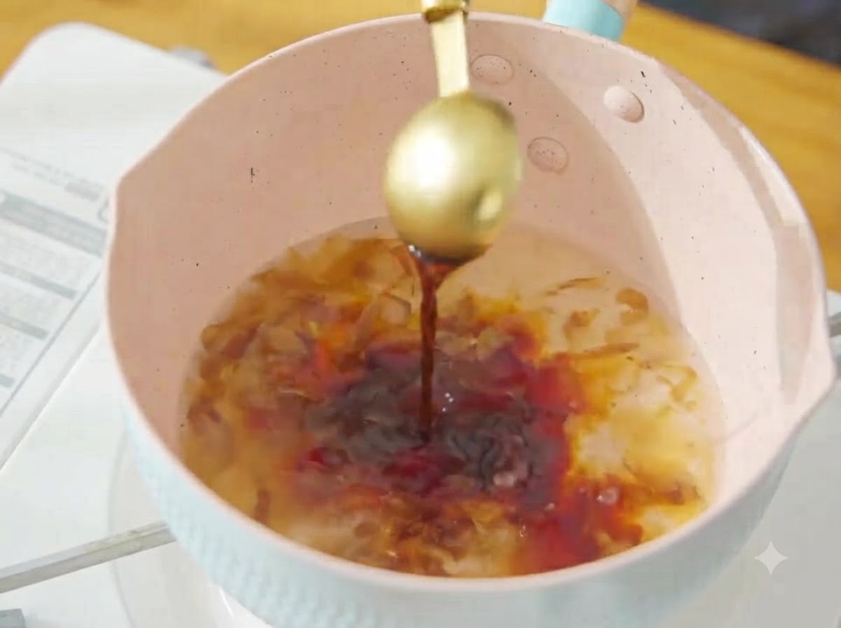 핑크색 냄비의 끓는 가쓰오부시 육수에 황금색 숟가락으로 진간장을 붓고 있는 모습.