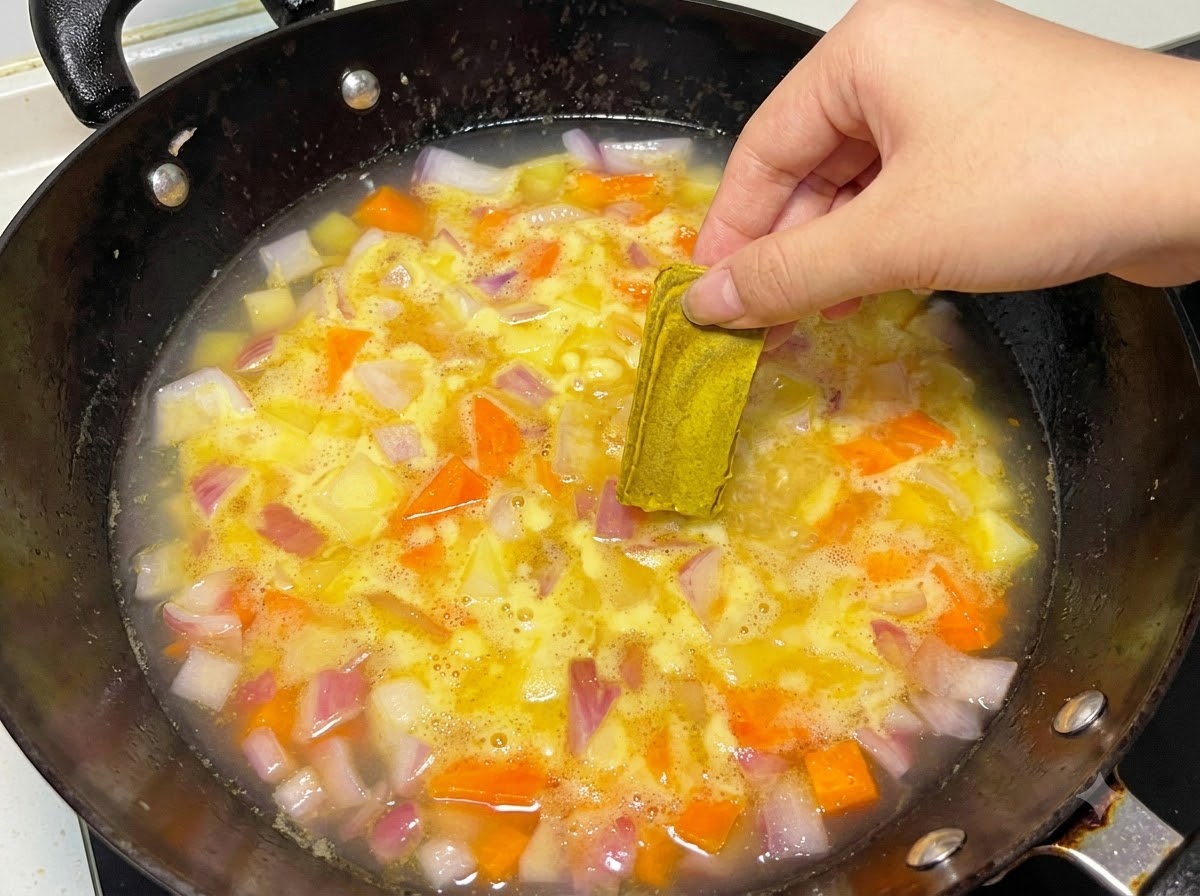 끓고 있는 물과 야채가 담긴 냄비에 손으로 고체 카레 블록을 넣는 모습.