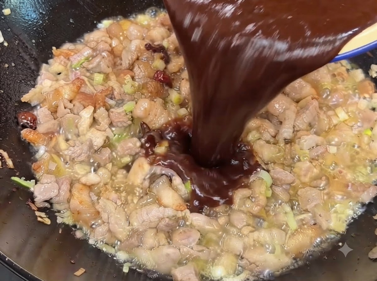 조리된 돼지고기 위의 웍에 어두운 된장 및 첨면장 소스 혼합물을 붓고 있는 모습.