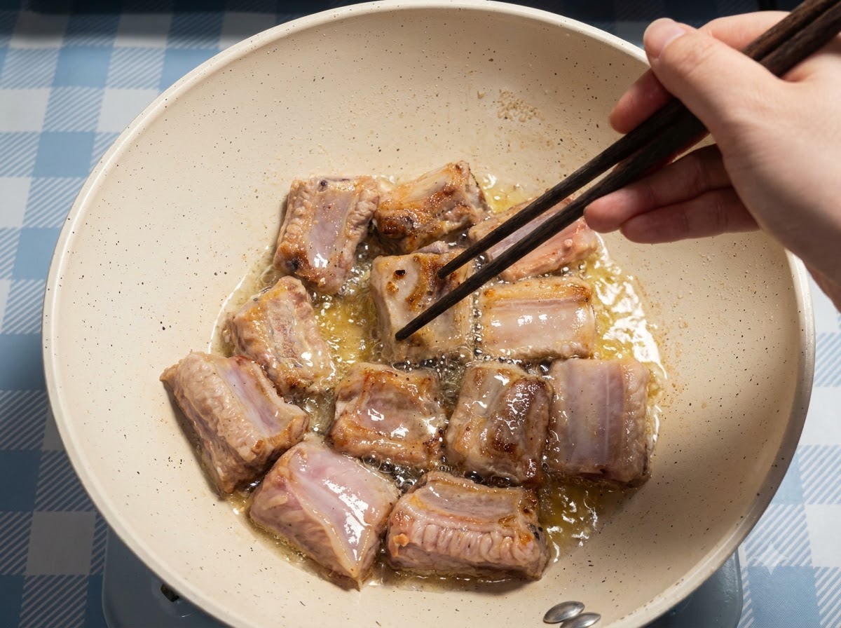 팬 안의 기름에서 여러 개의 돼지 갈비 조각이 튀겨지고 있으며, 손이 젓가락으로 이를 정리하고 있는 모습.
