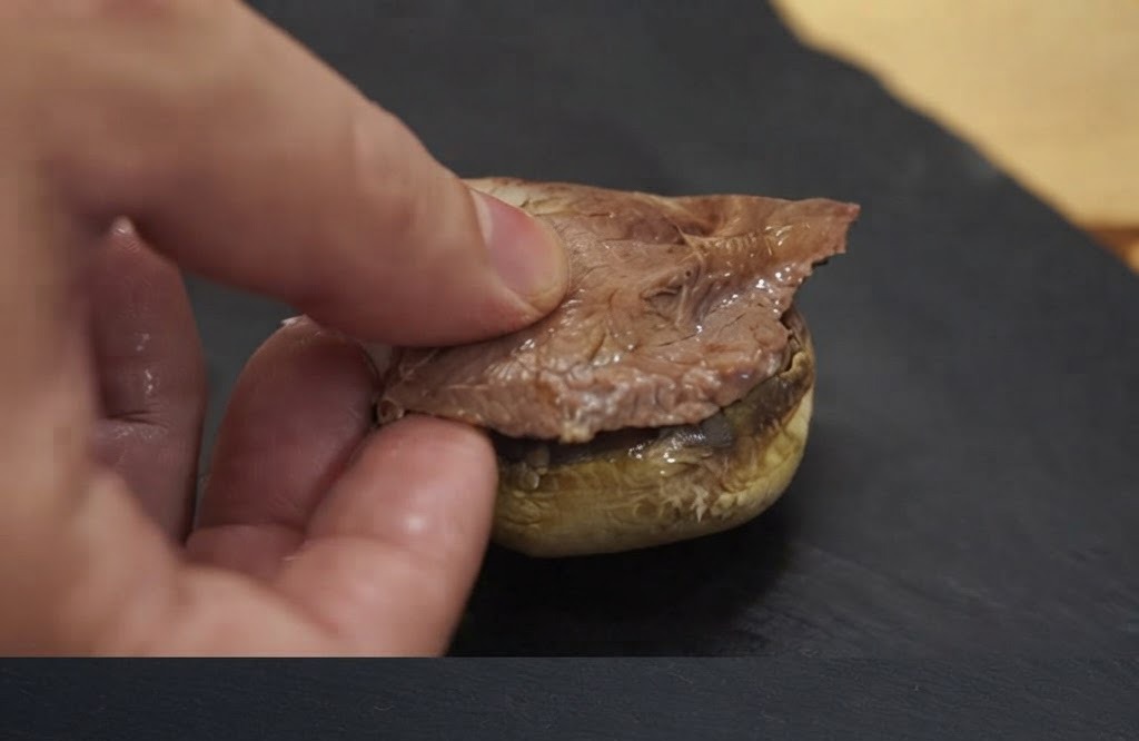 구운 버섯 캡 바닥 위에 조리된 고기 한 조각을 손으로 올리는 모습.
