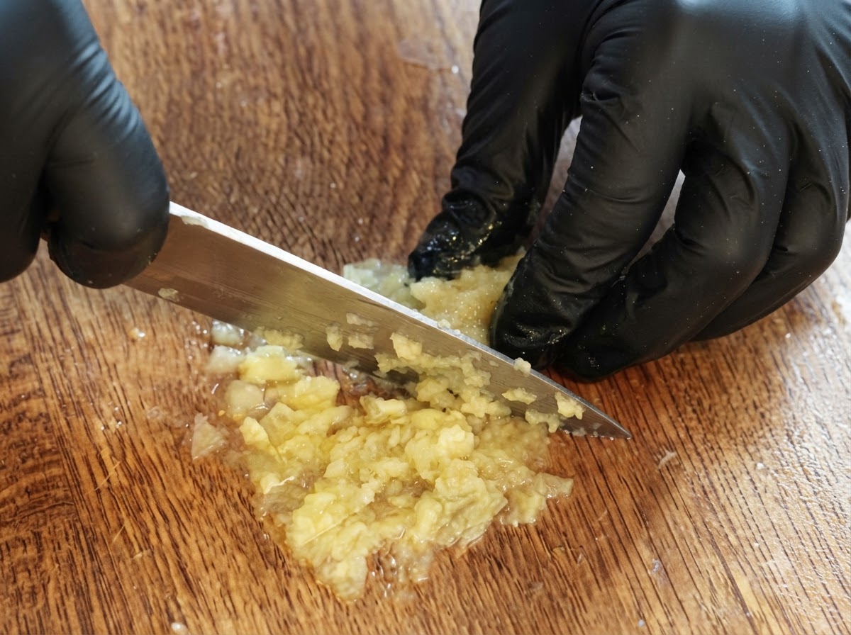 장갑을 낀 두 손이 나무 도마 위에서 남은 레몬 과육을 칼로 다지고 으깨어 퓨레로 만드는 모습