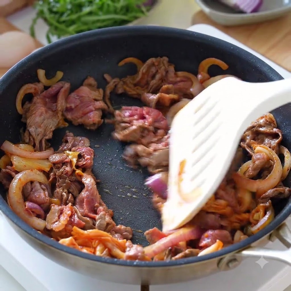 뒤집개를 이용해 프라이팬에서 썬 소고기, 양파, 김치가 함께 섞여 볶아지고 있는 모습.