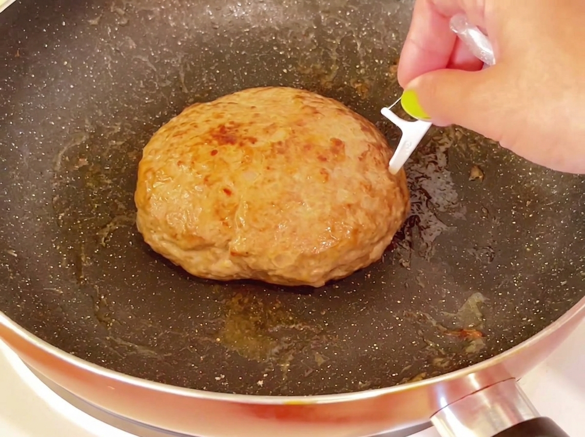 팬에 담긴 두툼하고 익은 햄버거 패티의 윗면에 이쑤시개를 찌르고 있는 모습.